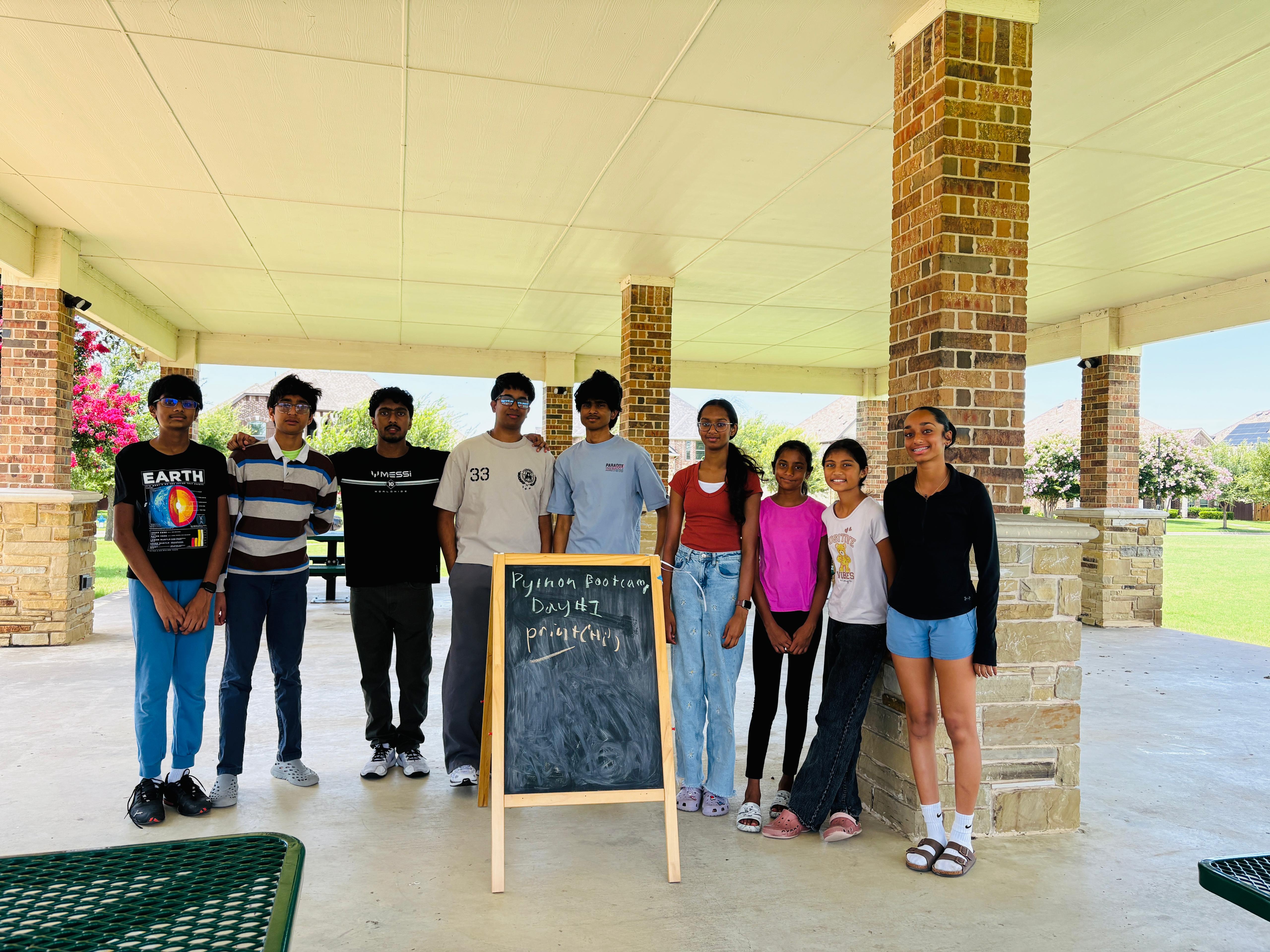 Students posing at an outdoor pavilion next to a chalkboard reading Python Bootcamp Day 1