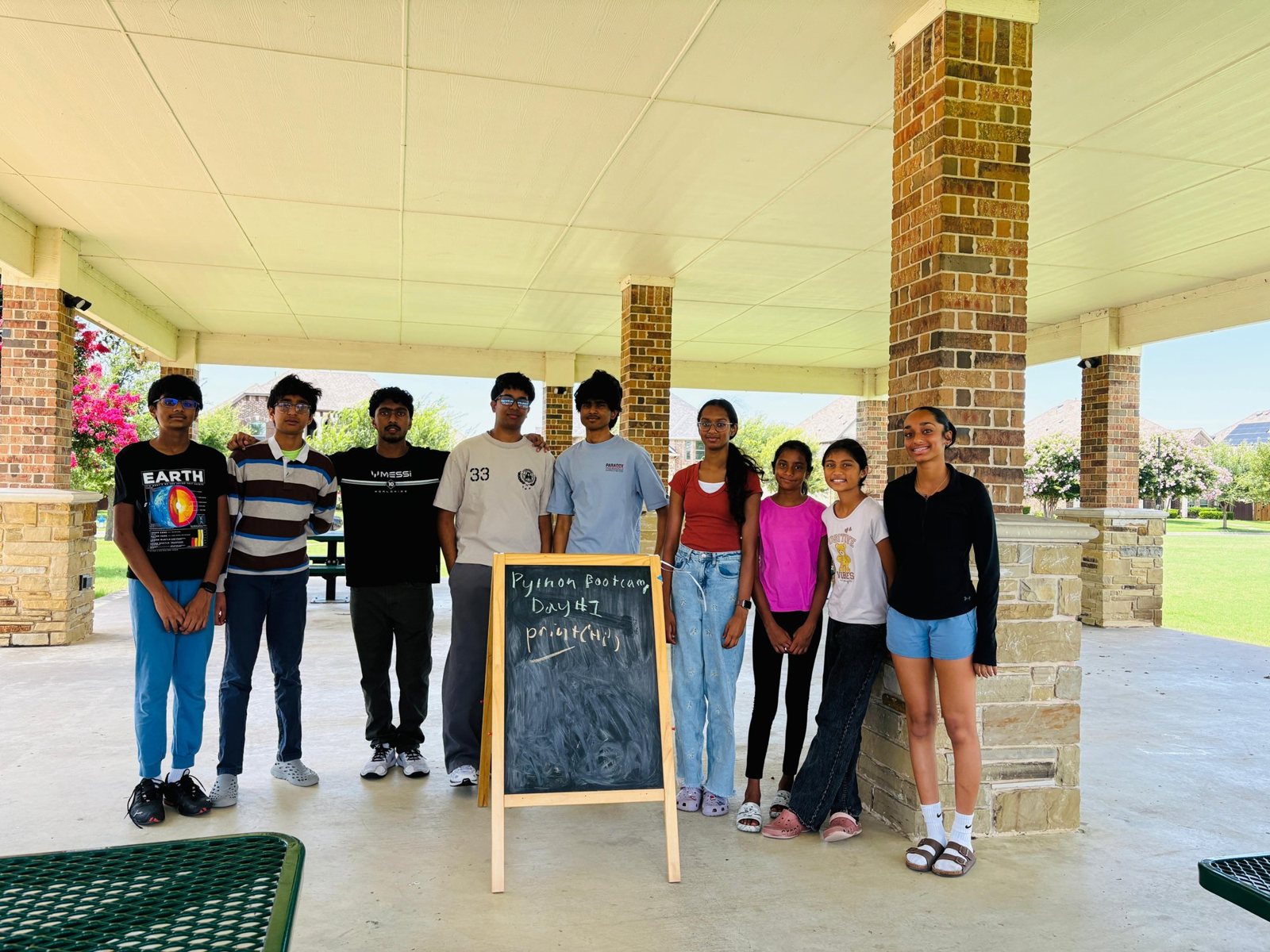 Nine people who helped run KidCode standing beside a chalkboard that reads Python Bootcamp Day 1, under an outdoor pavilion